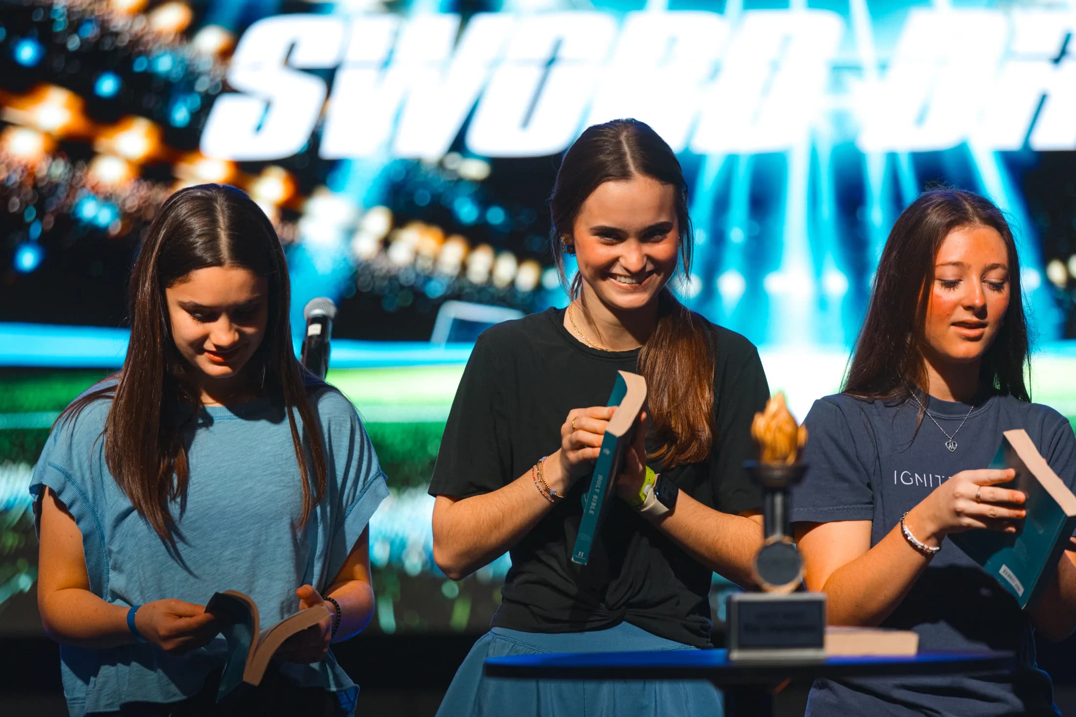 Three First Students participants hold open Bibles onstage beneath a large Students sign.